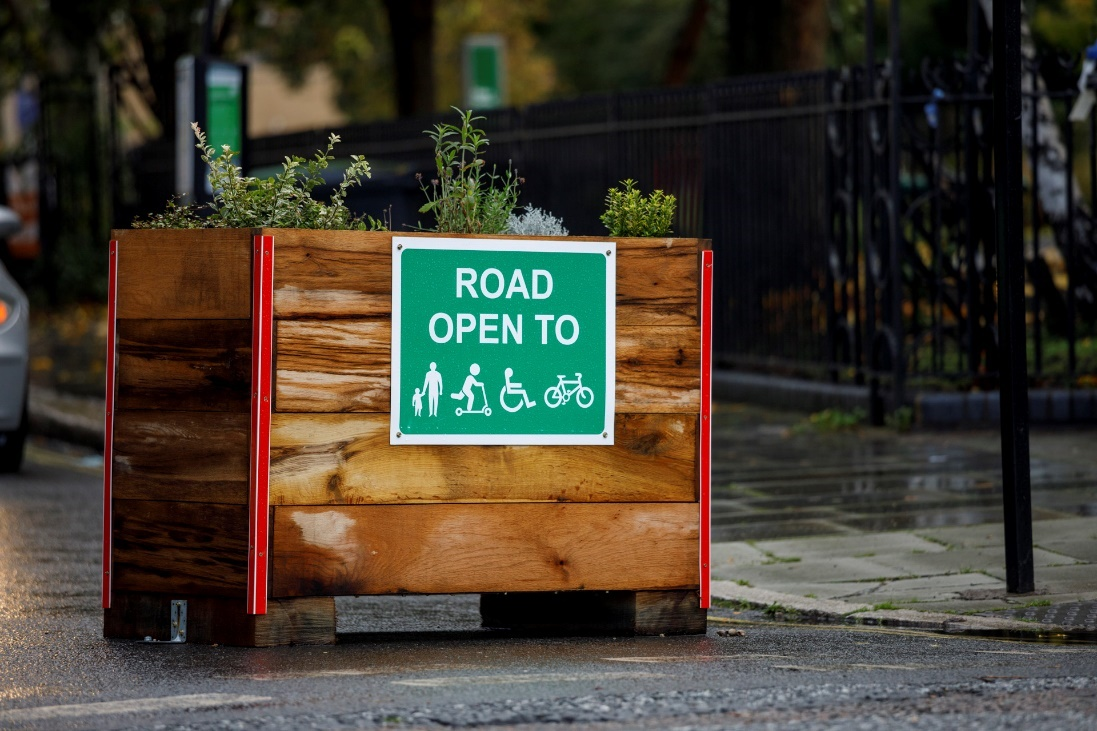 A wooden planter with some flowers in the middle of a residential street. On the front is a green sign saying 'ROAD OPEN TO', followed by icons representing an adult holding a child's hand, a person on a push scooter, a person using a wheelchair, a bicycle.